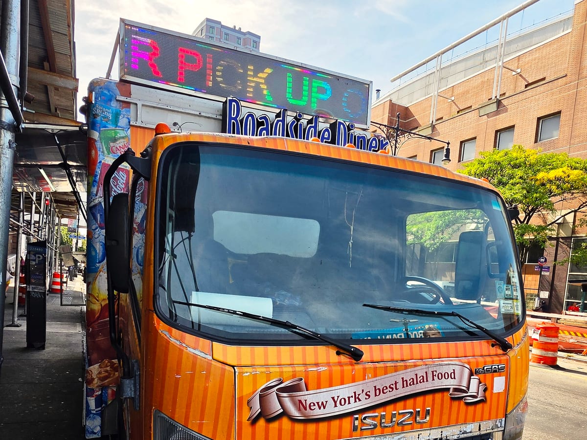 The front of the orange Roadside Diner truck on the Grand Concourse, with a lit PICK UP sign above and “New York’s best halal food” across the hood.